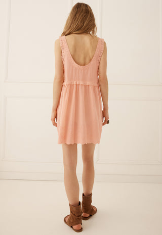 a-woman-viewed-from-behind-wearing-a-sleeveless-peach-pink-mini-dress-with-a-ruffled-scoop-neckline-and-brown-leather-slide-sandals-standing-against-a-white-paneled-wall