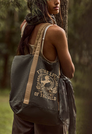 A woman outdoors by a tree wearing a dark olive canvas tote bag with beige straps and a distressed cream graphic that reads 'FOR THE LOVE OF THE WILD'.