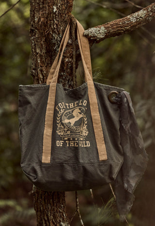 A dark gray canvas tote bag with tan straps and a distressed cream graphic reading 'FOR THE LOVE OF THE WILD' hanging from a tree branch in a forest setting.